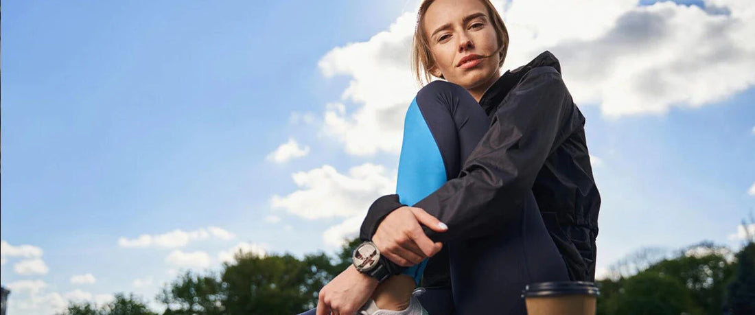 Junge Frau in Sportdress sitzt auf der Mauer, neben ihr ein To-Go-Becher Kaffee – Kaffee und Sport vereint.