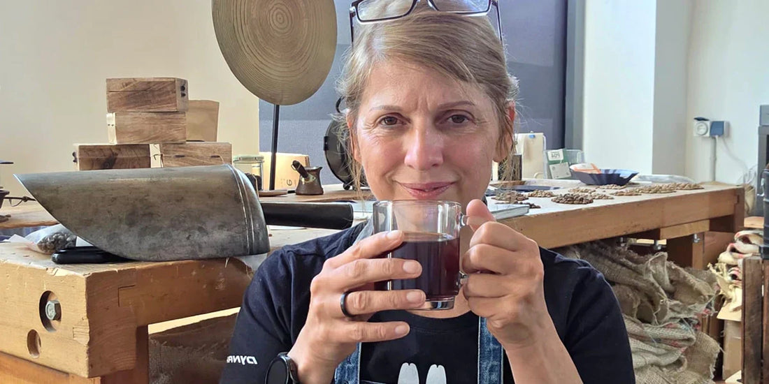 Tina mit Kaffeetasse in der Rösterei, Arbeitstisch im Hintergrund.