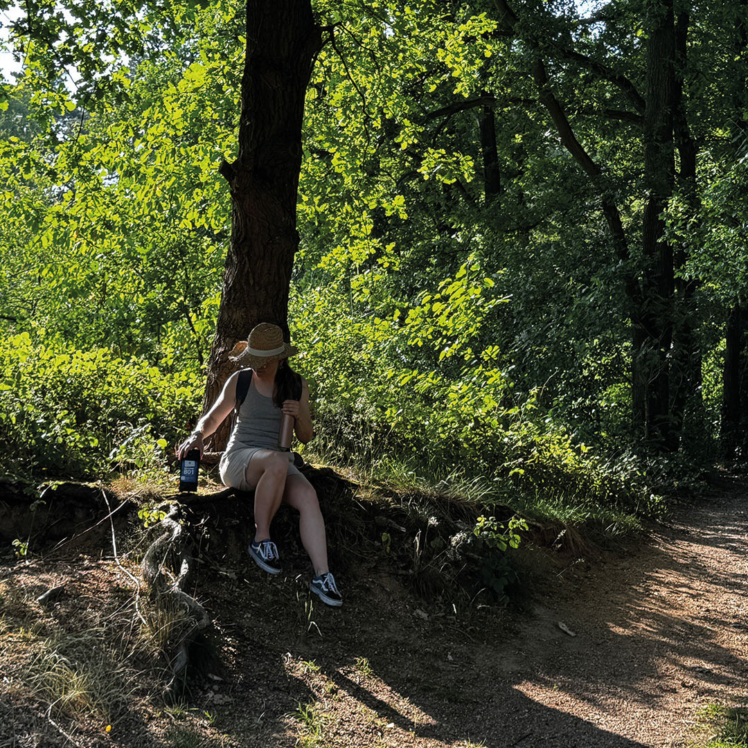 „Sandra macht eine kleine Pause auf einem Wanderweg, umgeben von schönem Grün. Mit einem Sonnenhut genießt sie die entspannte Atmosphäre und hat passend dazu den Wanderer, unseren leckeren Röstkaffee, dabei.“