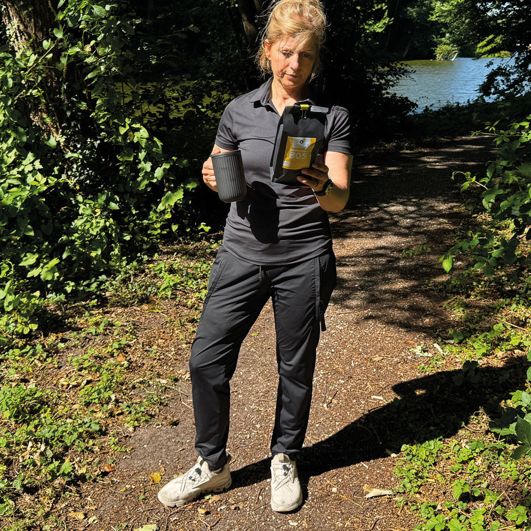 „Tina in einer wunderschönen, sonnigen Gegend mit einem kleinen See im Hintergrund. In der Hand hält sie eine Tasse und genießt den natürlichen Röstkaffee aus der schwarzen, gelben Verpackung – ein toller Filterkaffee, perfekt für diesen Moment.“