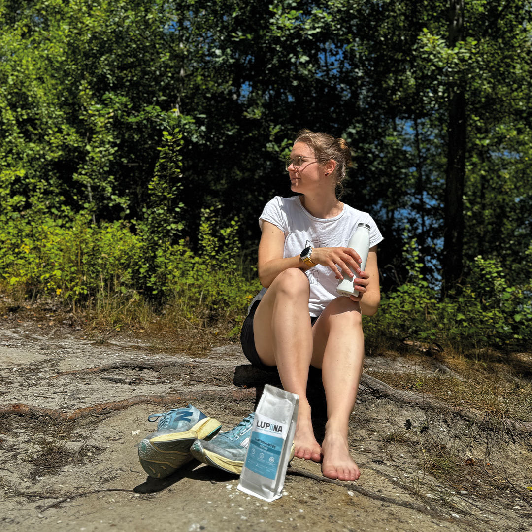Ein junges Mädchen sitzt entspannt in der Teverner Heide und genießt eine Pause nach einem langen Marsch. In der Hand hält sie eine himmelblaue Tasse mit Lupiina Activekaffee. Umgeben von natürlicher Heide-Landschaft strahlt die Szene Ruhe, Kraft und Erholung aus.