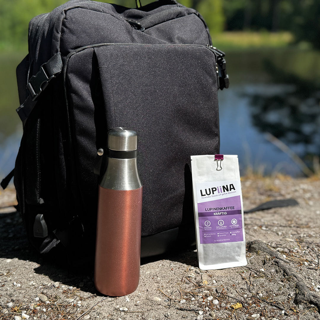 „Schwarzer Rucksack mit einer ToGo-Flasche Lupinenkaffee kräftig im Vordergrund, vor einem idyllischen See in der Teverner Heide.“