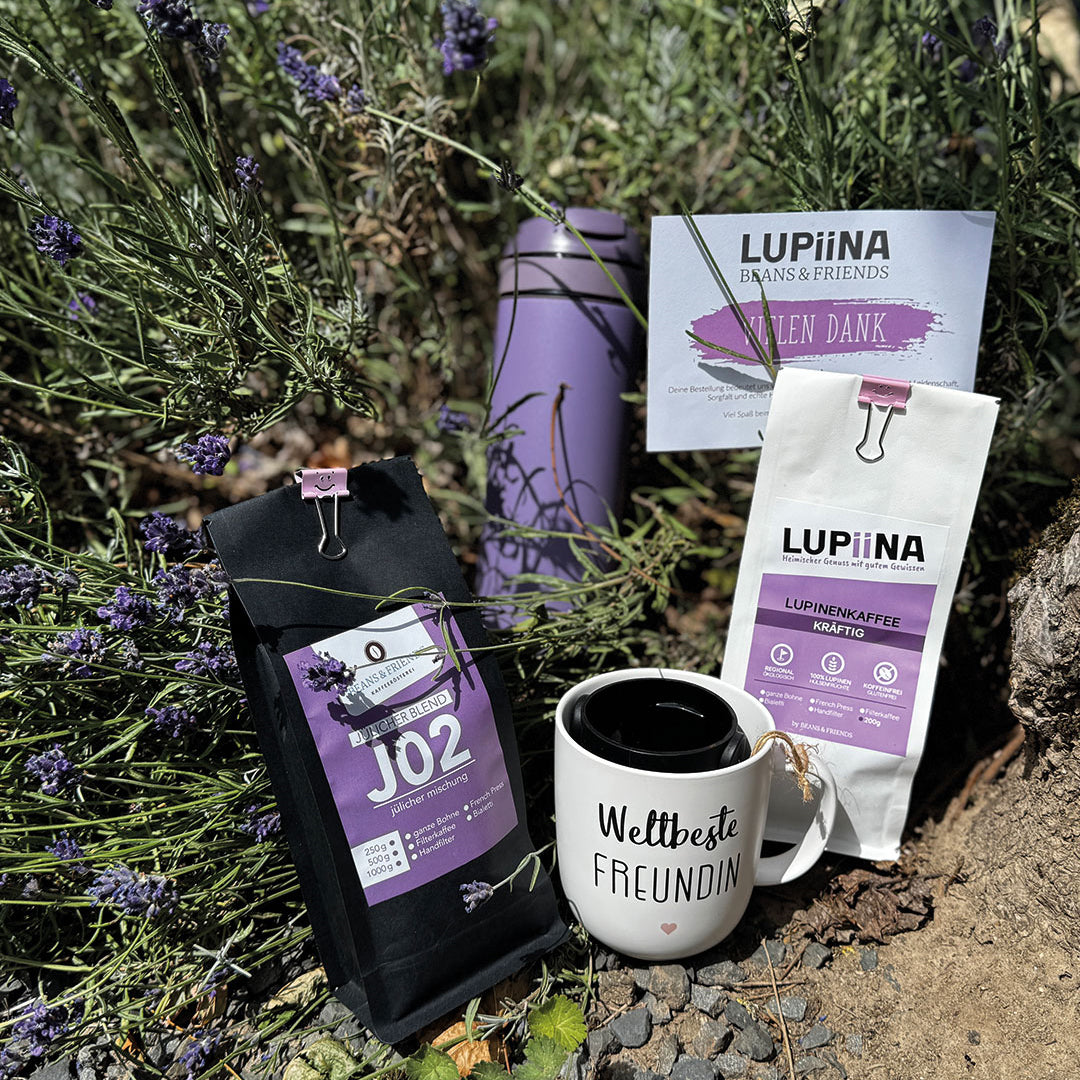 Zwei Kaffeesorten – Jülcher Mischung und kräftiger Lupinenkaffee – stilvoll in einem blühenden Lavendelfeld präsentiert. Daneben eine lavendelfarbene To-Go-Flasche und eine passende Tasse mit der Aufschrift „Beste Freundin“. Alles farblich harmonisch abgestimmt in Lavendel-Lila – ein Bild voller Ruhe, Genuss und Freundschaft.