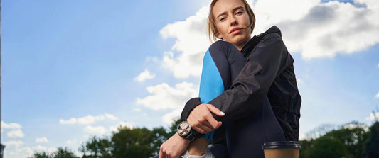 Junge Frau in Sportdress sitzt auf der Mauer, neben ihr ein To-Go-Becher Kaffee – Kaffee und Sport vereint.