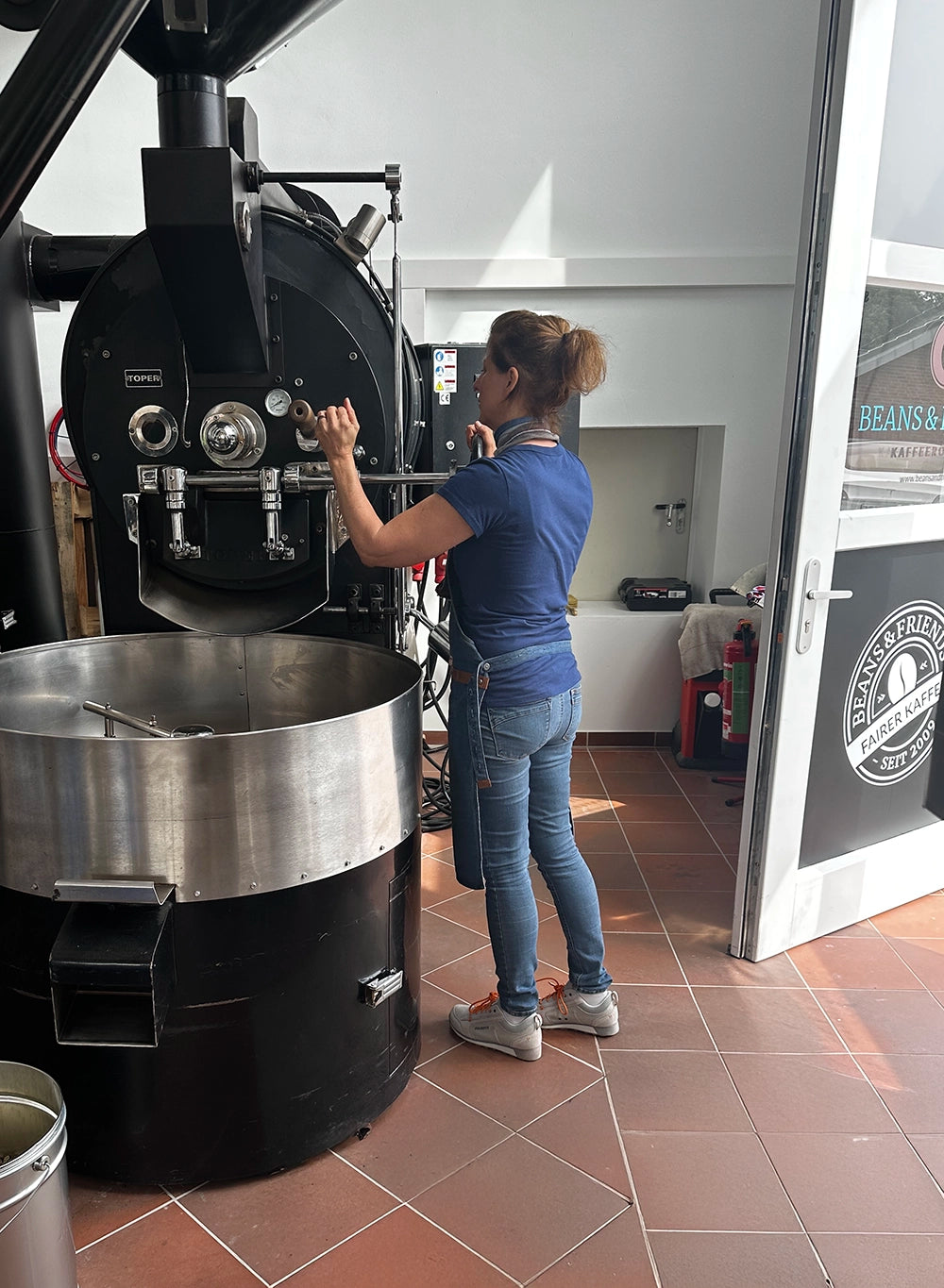 „Kaffeerösterin  Tina in ihrer Produktionshalle, zieht eine Probe während des Röstvorgangs an ihrem großen, schwarzen Röster – präzises Handwerk und Leidenschaft für Kaffeequalität."