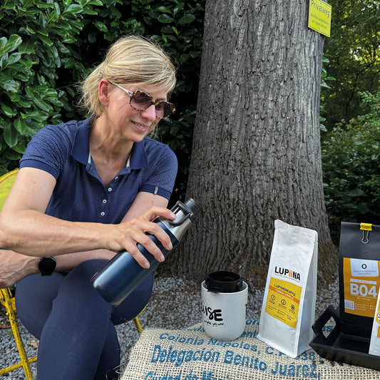 „Tina, in guter Stimmung und mit Sonnenbrille, hält eine blaue Isolierkanne in der Hand. Sie präsentiert den sonnigen Röstkaffee und den Lupinenkaffee Vanille, brüht lächelnd einen der beiden in einer Tasse auf.“

