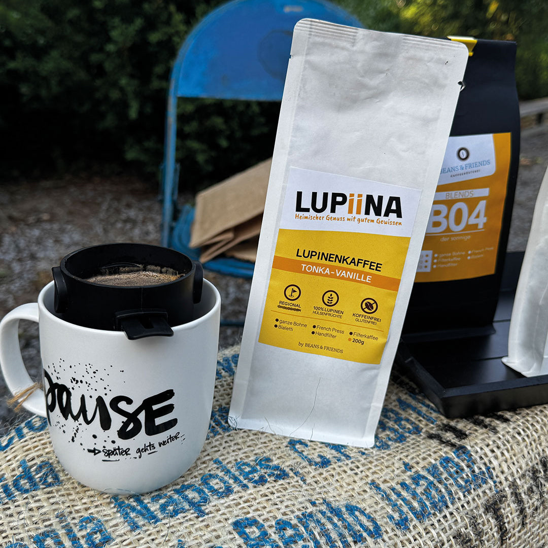 „Hier präsentieren wir den Lupinenkaffee Vanille Lupiina Tonka Vanille auf einem Kaffeesack. Daneben steht eine Tasse mit der Aufschrift ‚Pause‘, die das Bild hervorhebt und die entspannte Situation perfekt beschreibt.“
