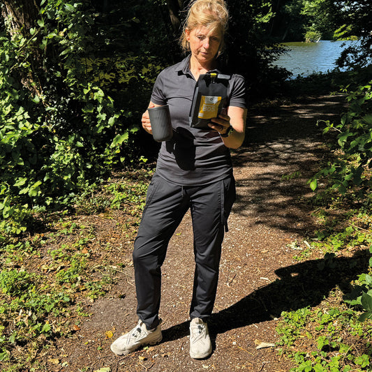 „Tina in einer wunderschönen, sonnigen Gegend mit einem kleinen See im Hintergrund. In der Hand hält sie eine Tasse und genießt den natürlichen Röstkaffee aus der schwarzen, gelben Verpackung – ein toller Filterkaffee, perfekt für diesen Moment.“

