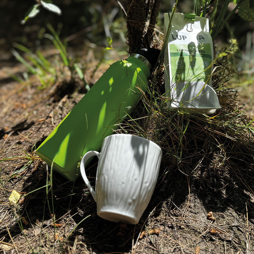 „Lupinenkaffee mild in weißer Verpackung mit grünem Etikett. Eine ToGo-Flasche und Tasse im Vordergrund, eingefangen im Sonnenlicht – harmonisches Zusammenspiel von Sonne und Schatten.“
