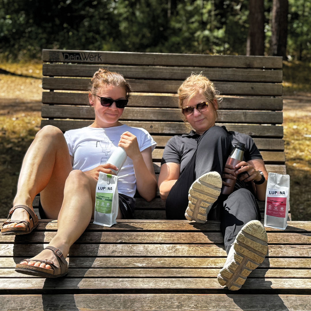 „Lupinenkaffee mild und Cranberry – Eine kleine Pause nach einer Wanderung in der Tevenrer Heide. Tina und Sandra genießen auf einer Bank einen Schluck ihres jeweiligen Lupinenkaffees aus der ToGo-Flasche.“
